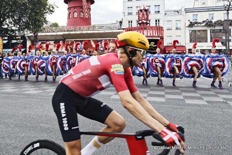 Un coureur du Tour de France passe devant le Moulin Rouge tandis que les danseuses du cabaret poursuivent leur spectacle place Blanche à Paris.