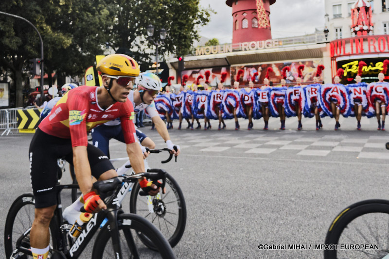 Des coureurs du Tour de France passent devant le Moulin Rouge tandis que les danseuses poursuivent leur spectacle place Blanche à Paris.