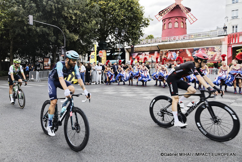 Plusieurs coureurs du Tour de France passent devant le Moulin Rouge tandis que les danseuses poursuivent leur spectacle place Blanche à Paris.