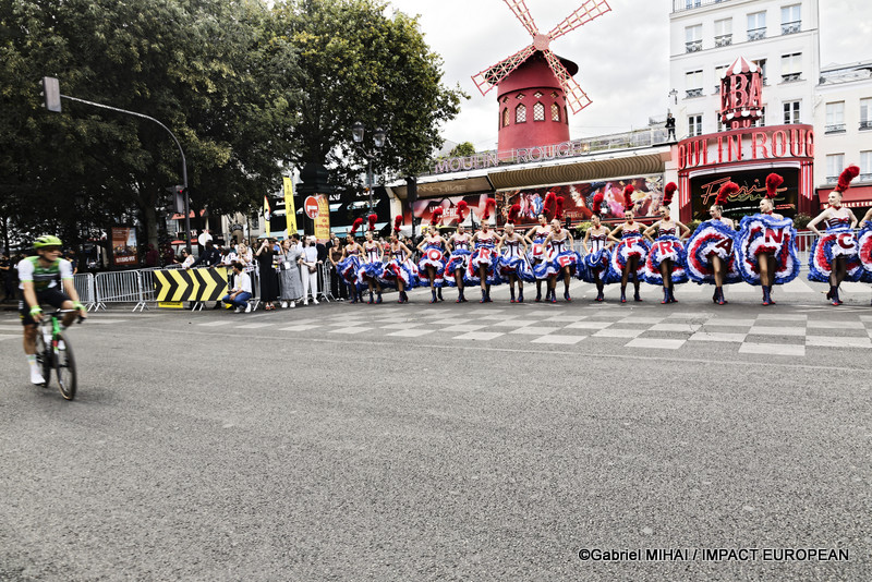Un coureur du Tour de France quitte la place Blanche tandis que les danseuses du Moulin Rouge poursuivent leur spectacle devant le célèbre cabaret parisien.
